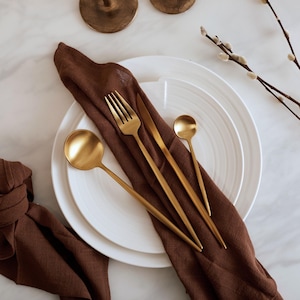 May include: A set of gold-toned cutlery, including a fork, knife, and two spoons, arranged on a brown napkin and white plate.