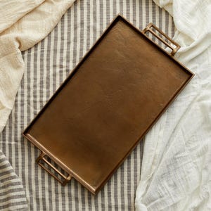 May include: A rectangular bronze serving tray with two rectangular handles. The tray is set on a striped grey and white fabric, with a cream-coloured fabric on the left and a white fabric on the right.