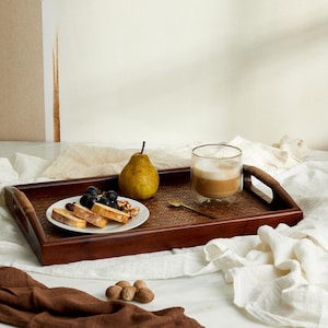May include: A wooden tray with a woven pattern, featuring a glass of cappuccino, a pear, grapes, and sliced bread. The tray is set on a white linen tablecloth.