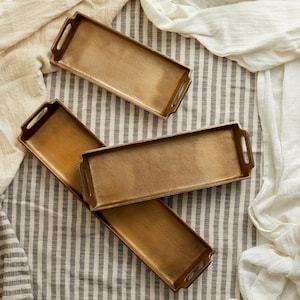 May include: Three gold-toned metal serving trays of different sizes, arranged on a striped fabric background.