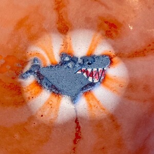 May include: A bath bomb with a shark design in a bath of orange water. The shark is gray with white teeth and a red mouth.