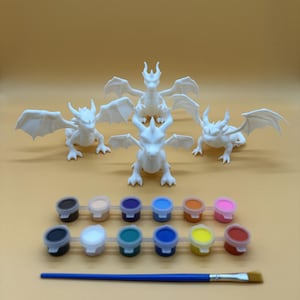 May include: Four white plastic dragon figurines with wings, ready to be painted. The set includes 12 paint pots in various colors, two paintbrushes, and a light brown background.