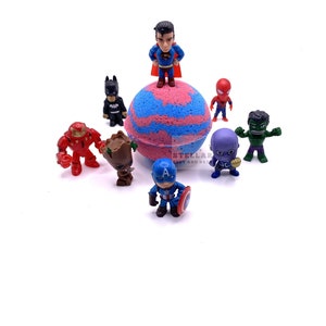 May include: A pink and blue bath bomb with a Superman figurine on top.  Surrounding the bath bomb are figurines of Batman, Iron Man, Groot, Captain America, Thanos, Spiderman, and the Hulk.