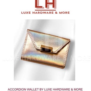 May include: A gold metallic accordion wallet with a black clasp. The wallet is from Luxe Hardware & More. The image includes the text "LH" and "LUXE HARDWARE & MORE" at the top. The text "ACCORDION WALLET BY LUXE HARDWARE & MORE" is at the bottom.