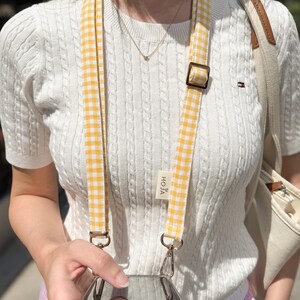 May include: A yellow and white gingham pattern phone strap with metal hardware. The strap is attached to a clear phone case. The word "HOJA" is printed on a small tag. The person is wearing a white cable knit sweater.