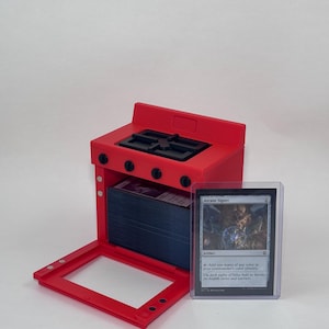 May include: A bright red plastic card storage box shaped like a miniature kitchen stove with an open oven door revealing a stack of trading cards inside. A single Arcane Signet trading card in a clear protective sleeve stands next to the box.