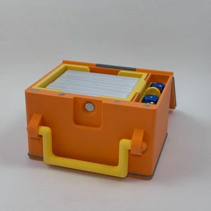 May include: An orange box with yellow accents, including a handle and interior trim. The box is open, revealing a stack of silver cards and a compartment with blue and yellow dice. The box is designed for storage.