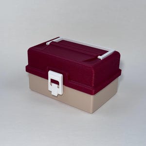 May include: A small, rectangular tackle box with a burgundy textured lid and a tan base. The box has a white handle and a white latch closure. The box is designed for storing small items.