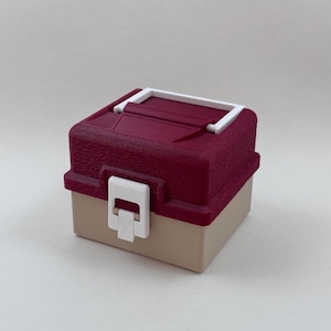 May include: A small, two-tone storage box with a burgundy textured lid and a tan base. The lid has a white accent and a white latch closure. The box is designed to be compact and portable.
