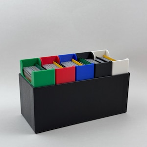 May include: A black card storage box with six colored dividers: green, red, blue, black, and white. Each divider holds a stack of cards. The box is designed to organize and store trading cards or other small items.