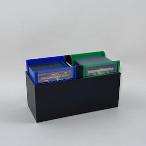 May include: A black card storage box with two compartments. Each compartment has a colored frame, one blue and one green, holding stacks of cards. The cards are visible, and text is present on some of the cards.