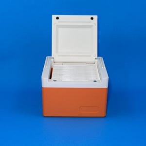 May include: An open, orange and white cooler on a blue background. The cooler has a white lid and a rectangular orange body. The interior of the cooler is visible, revealing a series of white, rectangular compartments. The cooler is designed to keep items cold.