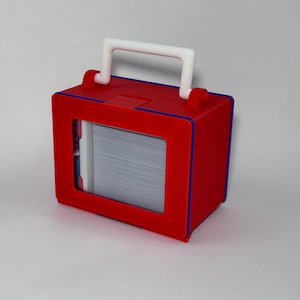 Könnte beinhalten: Eine rote Box mit weißem Griff und blauem Rand. Die Box hat ein rechteckiges Fenster, das einen Stapel Karten zeigt. Der Griff ist gewölbt und an der Oberseite der Box befestigt.