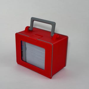 May include: A red rectangular box with a gray handle and a clear window displaying a stack of white cards. The box has gray trim and a latch on the top. The box is designed to hold cards.
