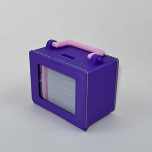 May include: A purple box with a pink handle and a window displaying a stack of white items. The box has a rectangular shape with a gray trim. The handle is arched and attached to the top of the box.