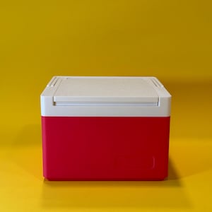 May include: A small, red and white cooler with a white lid. The cooler is sitting on a yellow surface.
