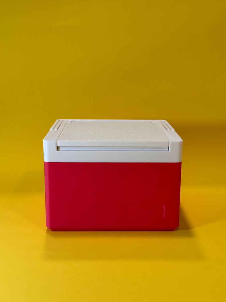 May include: A small, red and white cooler with a white lid. The cooler is sitting on a yellow surface.