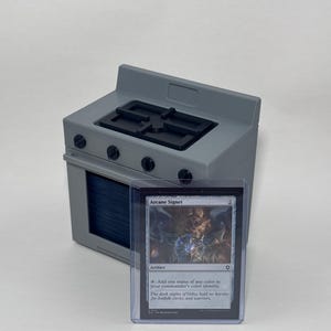 May include: A gray miniature stove with a black stovetop grate and four black control knobs, positioned behind a clear plastic protective sleeve containing a Magic: The Gathering trading card titled Arcane Signet with dark artwork.