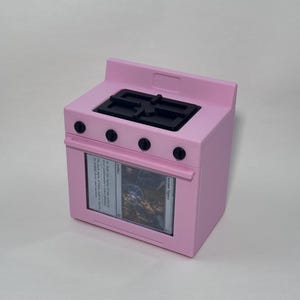 May include: A light pink toy stove featuring a black stovetop with four burners and four black control knobs. The oven door is a clear window displaying an Arcane Signet Magic the Gathering card inside, which includes text and artwork.