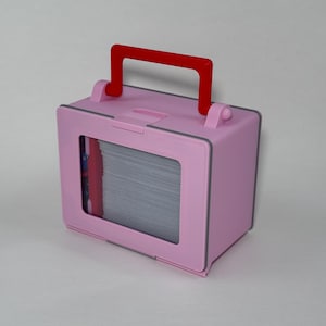 May include: A pink, rectangular storage box with a red handle. The box has a window on the front, revealing a stack of gray cards. The box has a gray trim and rounded corners. The handle is a bright red color.