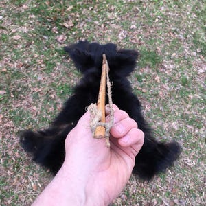 以下が含まれることがあります： 黒い毛皮の毛皮の上に、紐の付いた木製の道具を手が持っています。毛皮は草の上に広がっています。この道具は手作りのようです。