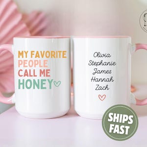 May include: White ceramic mug with a pink handle and rim. The front of the mug reads "MY FAVORITE PEOPLE CALL ME HONEY" in colorful text. The back of the mug has names and a heart symbol.