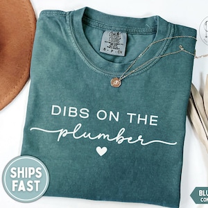 May include: Blue spruce Comfort Colors t-shirt with the phrase "DIBS ON THE plumber" in white script. A gold necklace with a pendant rests on the shirt. A brown hat and a palm frond are in the background.