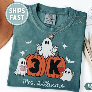 May include: A blue spruce Comfort Colors t-shirt featuring a graphic of three white ghosts and two orange pumpkins. One ghost holds a pink book, another holds a pencil, and the third makes a peace sign. The text reads 3K and Mrs. Williams.