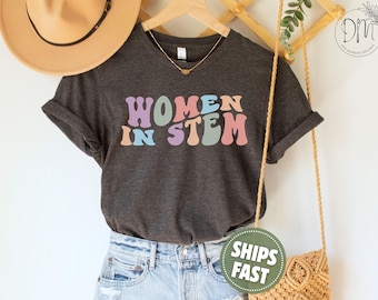 May include: A dark charcoal grey short-sleeve t-shirt featuring the text "WOMEN IN STEM" printed in a wavy, multi-coloured font with shades of purple, blue, peach, and green. The shirt is displayed on a hanger with a gold necklace.
