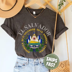 May include: A dark charcoal gray short-sleeve t-shirt featuring a graphic print of the El Salvador coat of arms in the center. The design includes the text EL SALVADOR and REPUBLICA DE EL SALVADOR EN LA AMERICA CENTRAL in a circular layout.