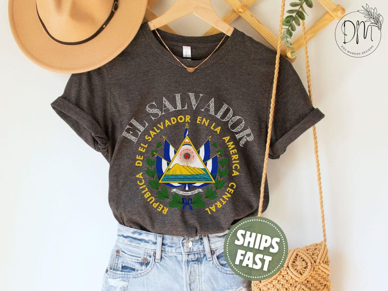 May include: A dark charcoal gray short-sleeve t-shirt featuring a graphic print of the El Salvador coat of arms in the center. The design includes the text EL SALVADOR and REPUBLICA DE EL SALVADOR EN LA AMERICA CENTRAL in a circular layout.