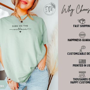 May include: A light green t-shirt with the text "DIBS ON THE villain" in black script. The shirt is part of the "Bay Comfort Colors" collection. The image also includes text about fast shipping, happiness guarantee, customizable designs, and being printed in the USA.