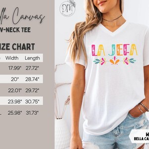 May include: White Bella Canvas V-neck tee with the text "LA JEFA" in colourful letters. The shirt has a size chart with measurements in inches. The tee features a floral design and is a casual wear item.
