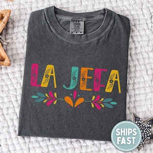 May include: Charcoal grey t-shirt featuring the words "LA JEFA" in vibrant pink, yellow, teal, and orange. The design includes floral and leaf motifs. The shirt is displayed on a textured white surface.