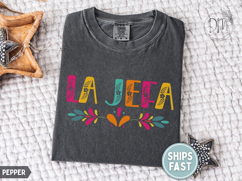 May include: Charcoal grey t-shirt featuring the words "LA JEFA" in vibrant pink, yellow, teal, and orange. The design includes floral and leaf motifs. The shirt is displayed on a textured white surface.