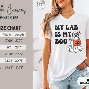 May include: A white V-neck t-shirt featuring a black graphic of a Labrador retriever wearing a ghost costume and holding an orange jack-o'-lantern bucket. The text reads MY LAB IS MY BOO. A size chart displays widths and lengths in centimetres.