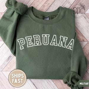 May include: Military green sweatshirt with the word "PERUANA" in white block letters. The sweatshirt has a crew neck and long sleeves. A round sticker with the words "SHIPS FAST" is on the bottom left. The words "MILITARY GREEN" are on the bottom right.