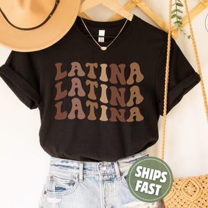 May include: A black short-sleeved t-shirt featuring the word LATINA printed three times in a stacked, wavy, retro-style font. The text appears in varying shades of brown, ranging from light tan to dark chocolate, set against the solid black fabric.