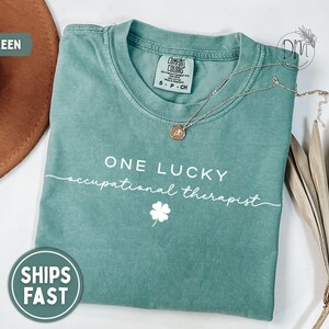 May include: Light green t-shirt with the text "ONE LUCKY occupational therapist" and a four-leaf clover graphic. A gold necklace rests on the shirt. The shirt is a Comfort Colors brand, size S-CH.