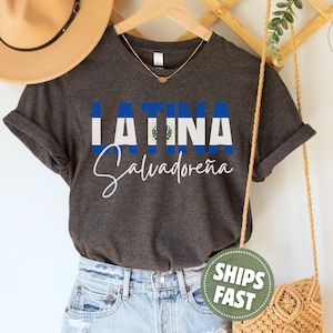 May include: A dark charcoal gray short-sleeve t-shirt featuring the text LATINA in block letters with the blue and white flag of El Salvador as the fill, and the word Salvadore&ntilde;a written in white cursive script underneath the flag text.