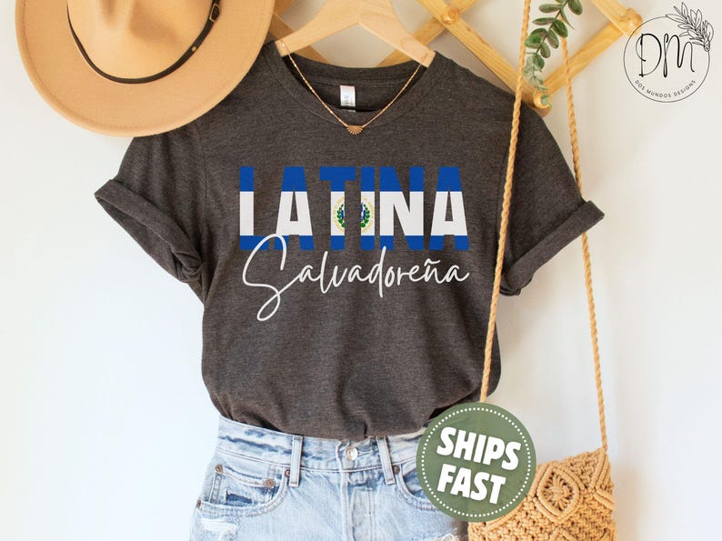 May include: A dark charcoal gray short-sleeve t-shirt featuring the text LATINA in block letters with the blue and white flag of El Salvador as the fill, and the word Salvadore&ntilde;a written in white cursive script underneath the flag text.