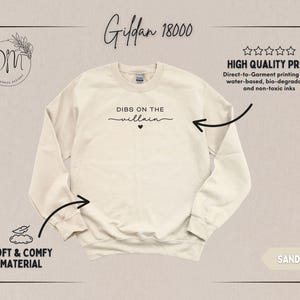 May include: Sand-colored Gildan 18000 sweatshirt with the phrase "DIBS ON THE villain" printed in black script. The sweatshirt features a crew neck and long sleeves. The image also highlights the soft and comfy material and high-quality print.