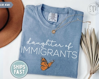 May include: A blue jean coloured t-shirt featuring the white text "daughter of IMMIGRANTS" and a graphic of an orange and black monarch butterfly. The shirt has a crew neckline and a Comfort Colors brand label visible at the inner collar.
