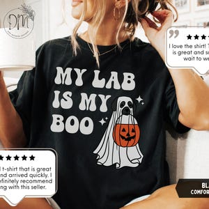 May include: A black t-shirt featuring the white text "MY LAB IS MY BOO" and a graphic of a Labrador dog wearing a white ghost costume while holding an orange jack-o'-lantern pumpkin bucket, accented with small white four-pointed star icons.