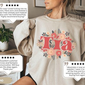 May include: Sand-colored sweatshirt with the word "Tia" in red, surrounded by a floral design in pink, peach, and blue. The sweatshirt has long sleeves and a crew neck. The word "SAND" is printed at the bottom.