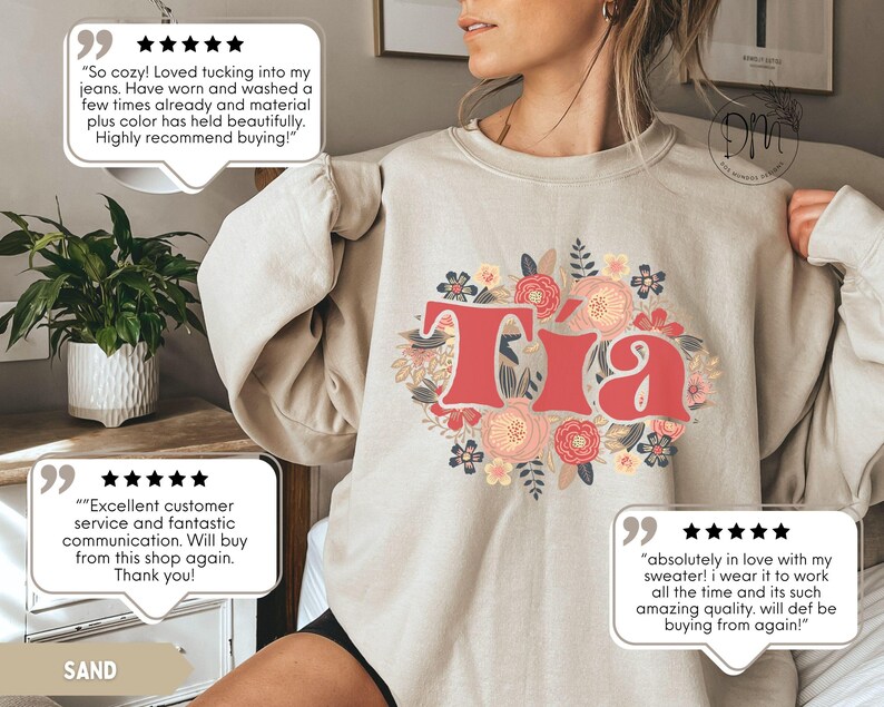May include: Sand-colored sweatshirt with the word "Tia" in red, surrounded by a floral design in pink, peach, and blue. The sweatshirt has long sleeves and a crew neck. The word "SAND" is printed at the bottom.