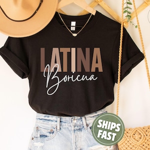 May include: A black short-sleeve t-shirt featuring the word LATINA in block letters with varying brown tones and the word Boricua written in white cursive script below it. The shirt has a crew neckline and is paired with light blue denim jeans.
