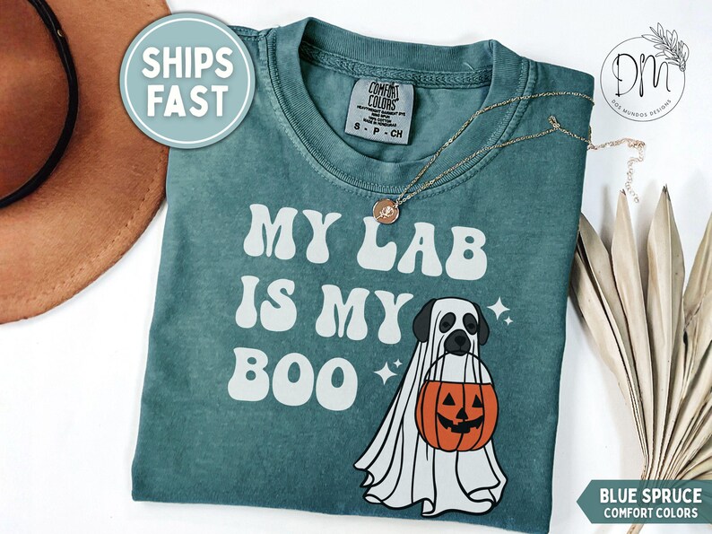 May include: A blue spruce Comfort Colors t-shirt featuring the white text MY LAB IS MY BOO and a graphic of a Labrador dog wearing a white ghost costume while holding an orange jack-o'-lantern pumpkin bucket with small white star accents.