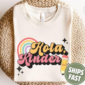 May include: A cream-colored short-sleeve t-shirt featuring a graphic print with the text "Hola, Kinder" in yellow and pink retro-style lettering. The design includes a colorful rainbow, black star accents, and a cartoon pencil character.
