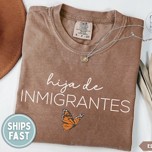 May include: A brown short-sleeved t-shirt featuring the white text "hija de INMIGRANTES" and a monarch butterfly graphic. The shirt has a Comfort Colors label at the neck and is displayed with a brown hat, a silver necklace, and dried palm leaves.
